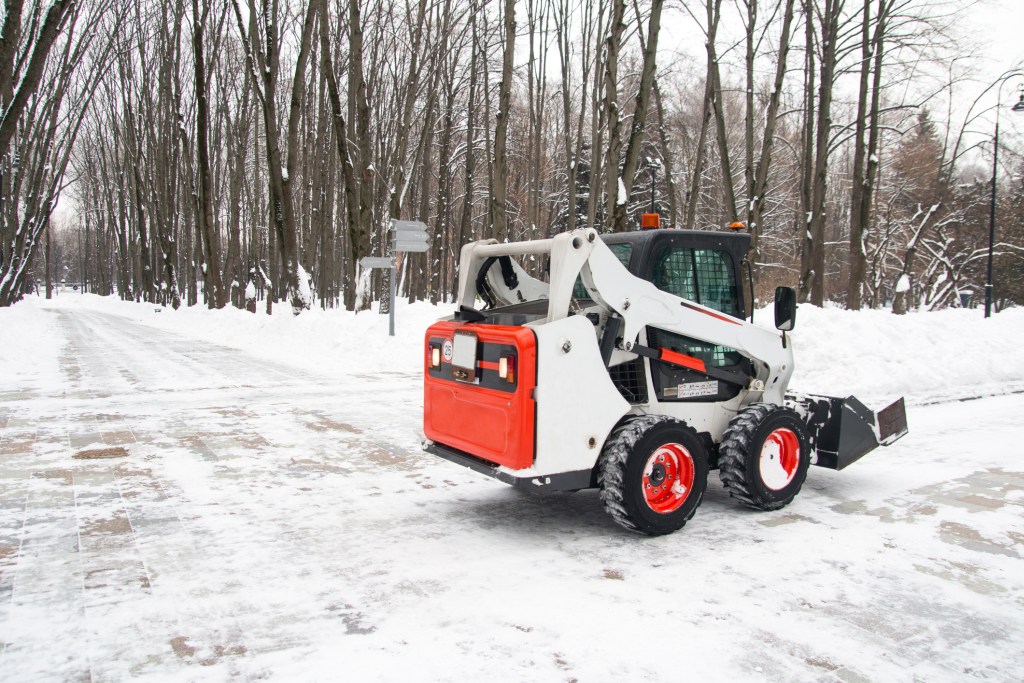 A bobcat removes snow on the road in a treed neighbourhood.
